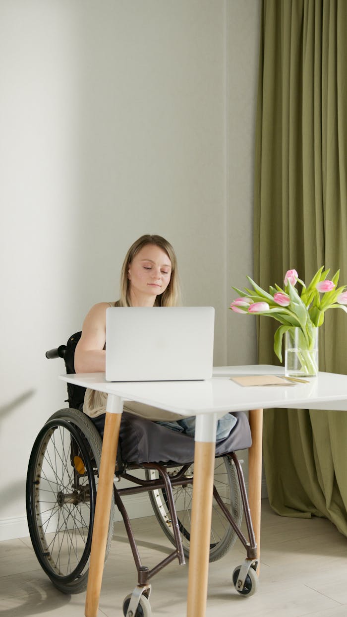 Woman in a wheelchair using a laptop at a table, indoors, near tulips.